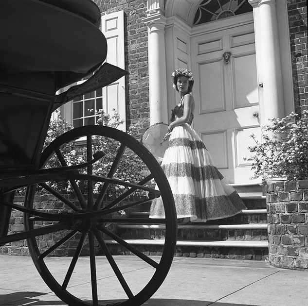 Госпожа Стэнли Мортимер позирует с зонтиком в вечернем платье на ступеньках дома в Чарльстоне, штат Южная Каролина. На переднем плане — очертания колеса экипажа.