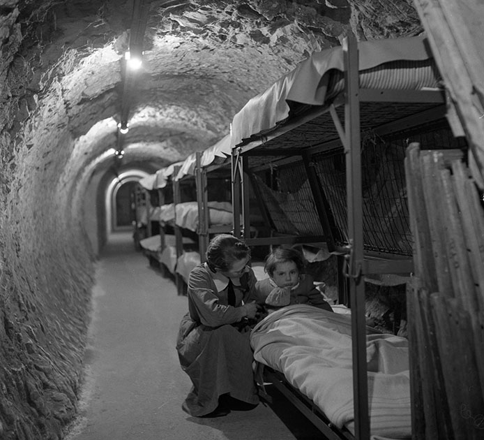 Женщина встает на колени рядом с кроватью ребенка в подземном тоннеле. Бомбежки Лондона во время Второй мировой войны, январь 1945 года.