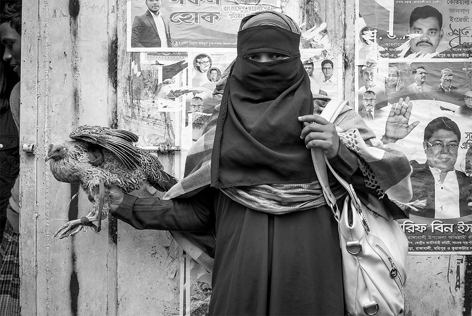 Фотография была сделана ранним утром в одном из людных районов Дакки, столицы Бангладеш. Художник был очарован прекрасно одетой женщиной, борющейся с только что купленной на рынке живой курицей. Он запечатлел ее закутанную фигуру на фоне стены, с которой на зрителей смотрят мужчины, столь контрастирующие с героиней снимка.