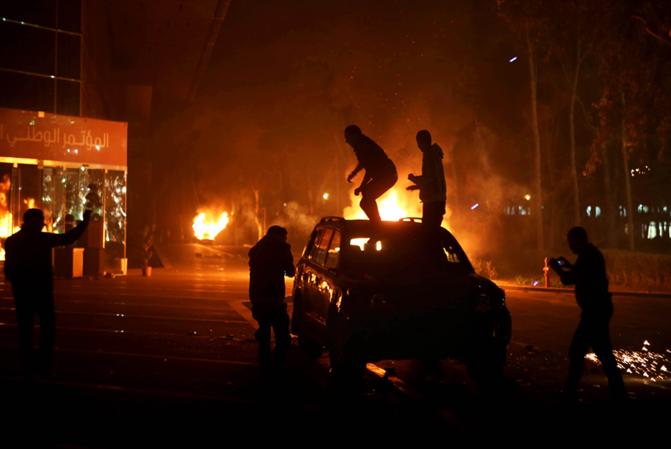 Демонстранты на фоне огня взбираются на автомобиль, припаркованный у здания Национального конференц-зала. Протестующие и вооруженные мятежники заставили временное правительство спешно покинуть это место.