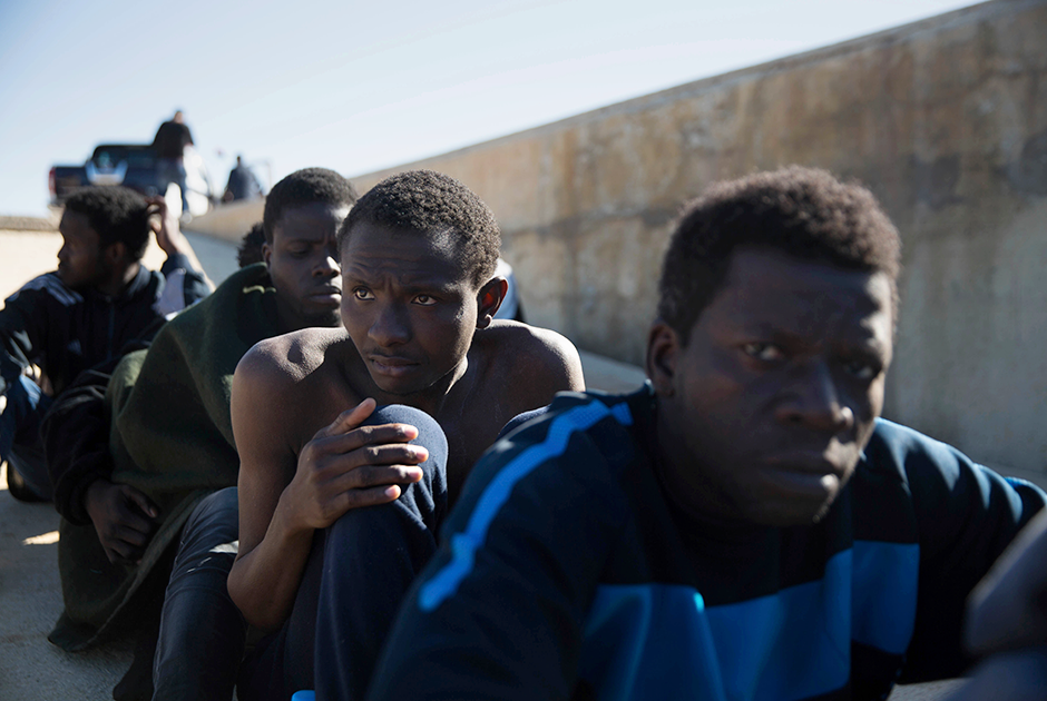 Мигранты отдыхают на тротуаре в порту. Они были спасены береговой охраной у побережья Триполи.