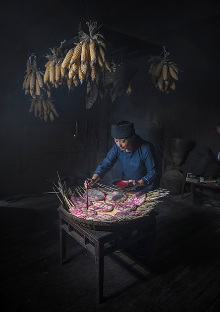 «Рисуя счастье» авторства Мин Кай Чан. Снимок сделан в удаленной деревне в западной части провинции Хунань в Китае, где проживает этническое меньшинство. Осенью люди делают заготовки еды на зиму. Деревенская женщина рисует традиционные символы на сухом рисовом пироге в надежде на счастливый и здоровый год.   

