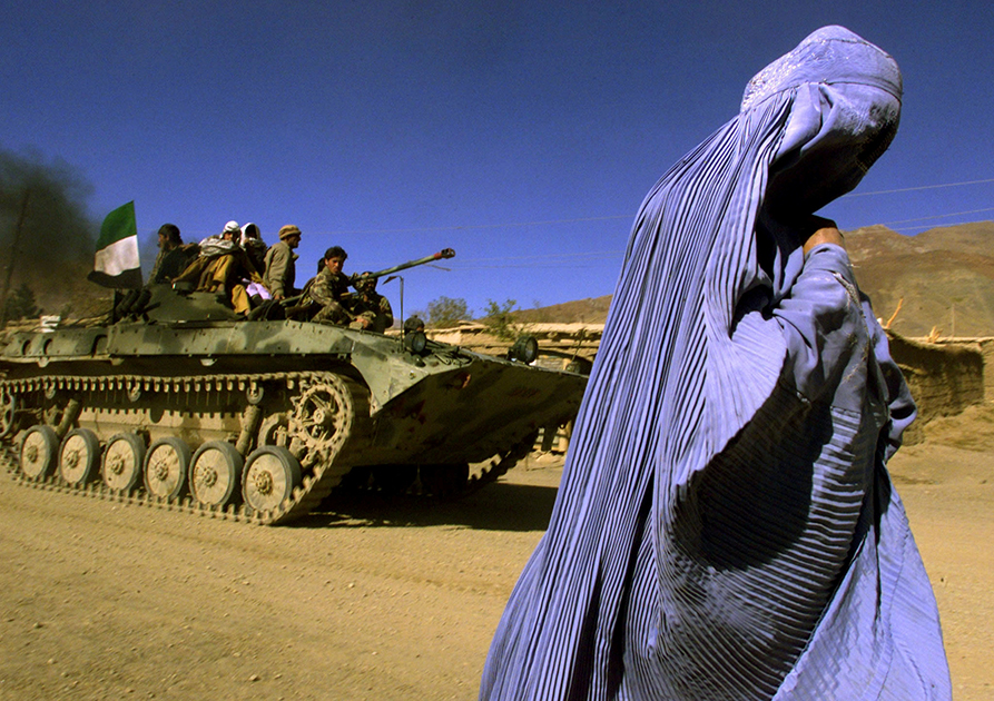 Жительница Афганистана в традиционной парандже идет по обочине. Рядом с ней — броневой транспортер Объединенного исламского фронта спасения Афганистана (организация также известна как «Северный Альянс»), украшенный флагом. Вооруженные бойцы выезжают на новую позицию по дороге  Кабул — Джабель — ус-Серадж. Снимок сделан 4 ноября 2001 года.