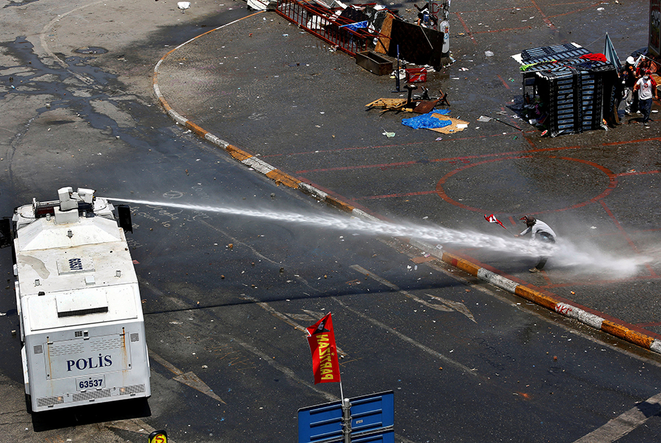 Протестующий падает под напором водяной пушки, в то время как его соратники прячутся за импровизированным укрытием во время столкновений на площади Таксим в Стамбуле. В июне 2013 года местные военные применили слезоточивый газ и водометы к сотням демонстрантов, которые пытались вернуть под свой контроль центр Стамбула.