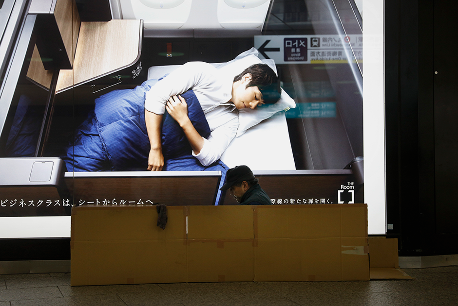Бездомный готовится ночевать на станции Синдзюку возле экрана с рекламой