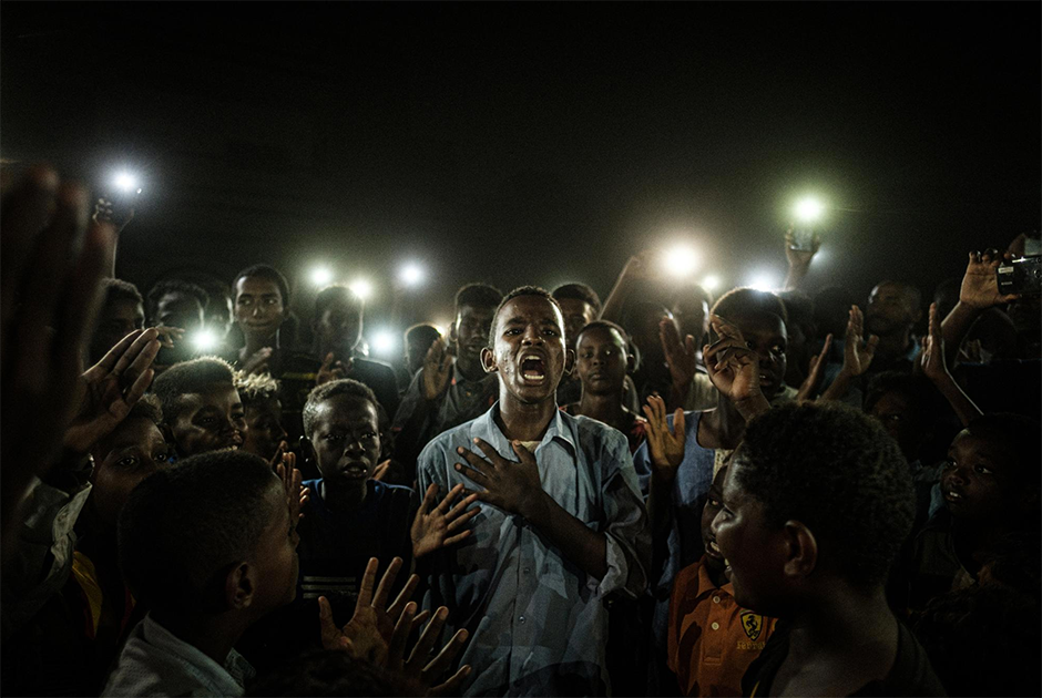 Юноша в свете мобильных телефонов декламирует стихотворение. Рядом с ним протестующие скандируют лозунги с требованиями перехода страны к гражданской форме правления. Снимок, представленный в номинации «Фотография года», был сделан 19 июня 2019 года в столице Судана Хартуме во время отключения электричества. Отключив свет и интернет, местные власти намеревались остановить митингующих.


Протесты начались на востоке страны в декабре 2018 года в ответ на ухудшение уровня жизни на всех уровнях. Предпосылкой стало повышение цен на хлеб в три раза. Вскоре демонстрации распространились по всей стране. Протестующие требовали отставки Омара аль-Башира — он был президентом Судана с 1993 года. 11 апреля в результате военного переворота его свергли и арестовали, а власть оказалась в руках Переходного военного совета. Протесты продолжились, и 3 июня правительственные войска открыли огонь по безоружным демонстрантам. По данным оппозиции, погибло более 100 человек. 