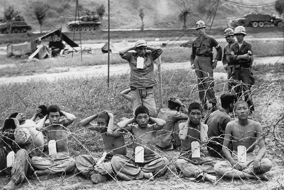 Военная полиция США охраняет северокорейских солдат, захваченных в плен 4 сентября 1950 года на линии фронта у реки Нактонган, во временном лагере для военнопленных недалеко от города Сувона. На заднем плане видно передвижение американских танков. 