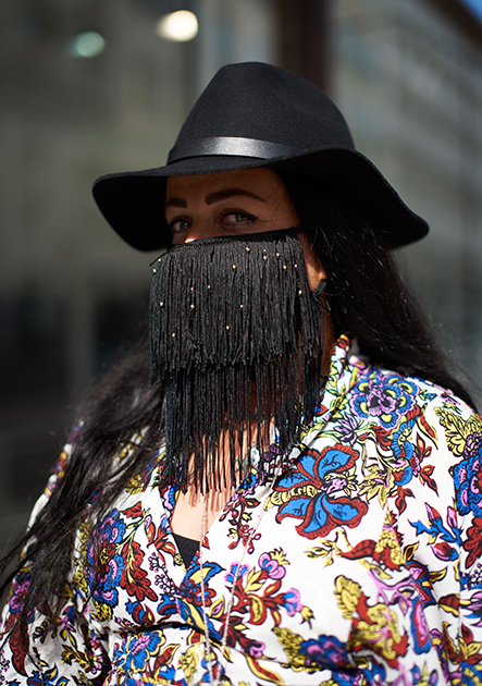 Одна из посетительниц лондонской Недели моды создала образ с довольно необычными смешениями цветов и стилей. Черная шляпа с кожаным обрамлением и маска для лица, украшенная бахромой, в сочетании с рубашкой с цветочным принтом навеяны Диким Западом в сочетании с гавайской культурой.

