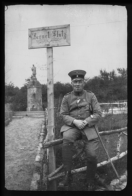 Немецкий офицер с «Железным крестом» и сигарой сидит на заборе возле таблички Бернет Платц. Западный фронт, 1916 год.

