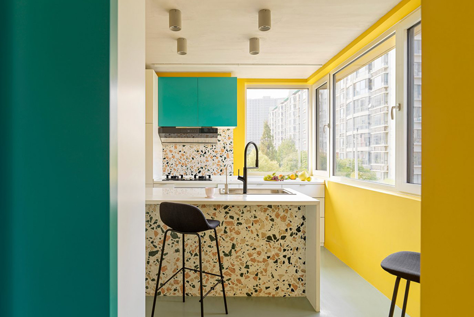 Архитекторы из китайского бюро MDDM Studio придумали яркий интерьер для кухни в пекинской квартире и выкрасили часть стен в желтый цвет. Несмотря на небольшое пространство, они смогли разместить в комнате и четырехконфорочную плиту, и столешницу с рабочей зоной и барной стойкой. Большая часть поверхностей — белая и глянцевая, но есть и яркие элементы в виде бирюзового шкафа и темно-серых барных стульев из текстиля.Цвета из буйной палитры сочетаются между собой. Дизайнеры достигают гармонии с помощью каменных панелей тераццо, которые изначально использовались на полах в венецианских интерьерах. На кухне они выделяются под барной стойкой и на стене около плиты, но интерьер не смотрится аляпистым — тераццо соединяет более теплые оттенки главных цветов комнаты, органично создавая единую концепцию.