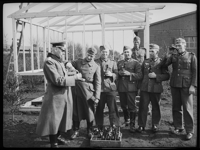 Немецкие солдаты и офицер угощаются алкоголем возле бараков для военнопленных в шталаге Зандбостель. Германия, август 1940 — июль 1941 года.