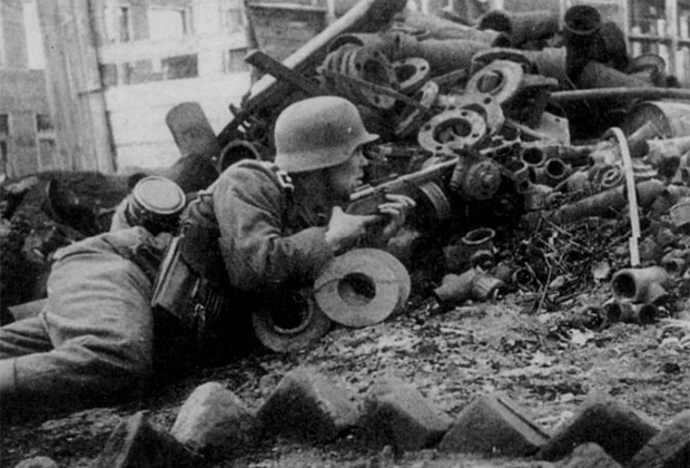 Немецкий унтер-офицер с трофейным советским пистолетом-пулеметом ППШ прячется за кучей металлоизделий во дворе завода. Сталинград, сентябрь-октябрь 1942 года