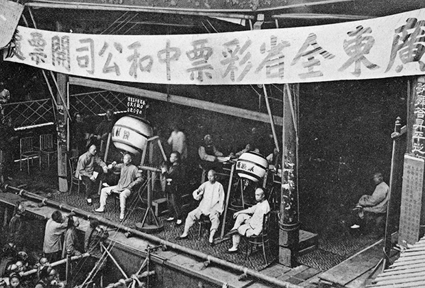 Engraved postcard of a group of men sitting on a stage drawing lottery numbers from raffle drums, with the lottery players looking on, Canton, also known as Guangzhou, Guangdong province, China, published by M. Sternberg, 1910. From the New York Public Library. (Photo by Smith Collection/Gado/Getty Images)