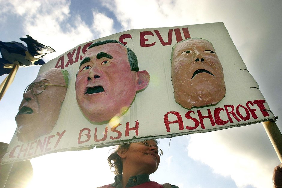 Женщина протестует против вторжения в Ирак, держа плакат с масками Джорджа Буша-младшего, вице-президента Дика Чейни и генпрокурора Джона Эшкрофта
