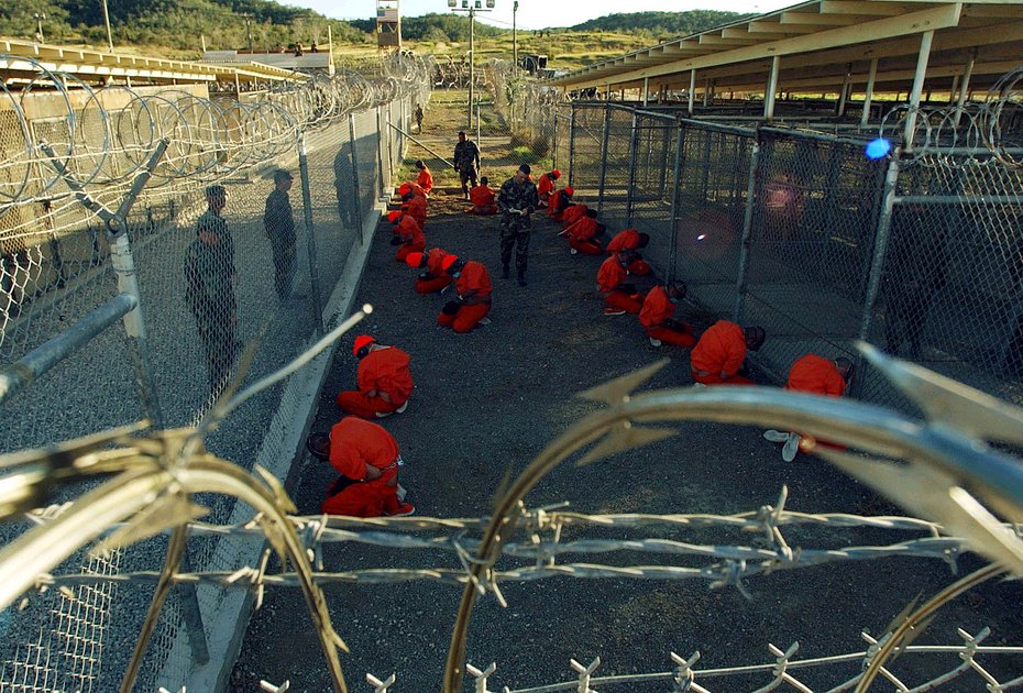 Заключенные в оранжевых комбинезонах сидят под бдительным присмотром военной полиции в лагере X-Ray на военно-морской базе Гуантанамо, Куба, 11 января 2002 года