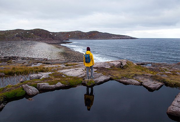 Tourist girl in yellow raincoat with backpack standing on shore of Barents Sea, seagulls fling over pebble beach. Teriberka, Murmansk Oblast, Russia